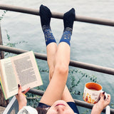 Person sitting by a body of water with legs crossed, wearing blue socks and holding a book and mug. thumbnail