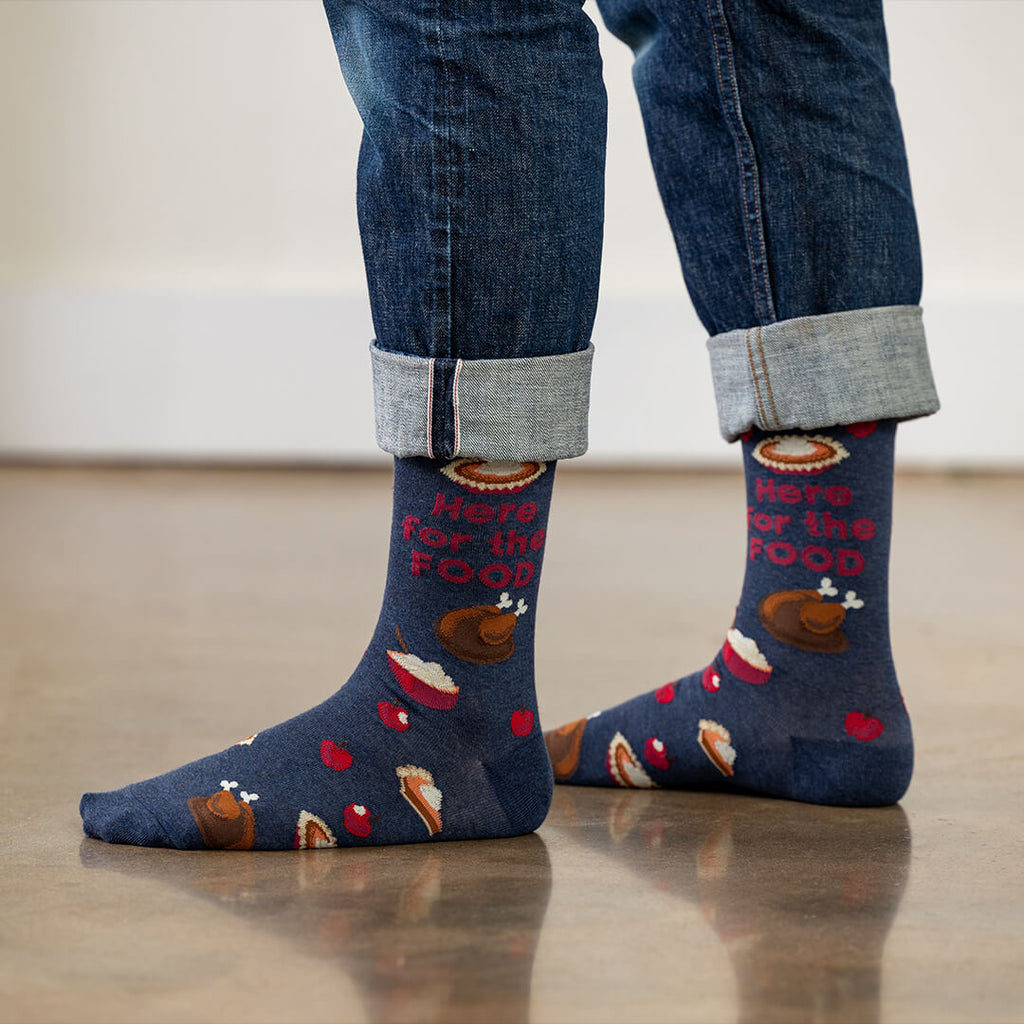 Person wearing blue socks with Thanksgiving-themed design and rolled-up jeans on a light wooden floor.