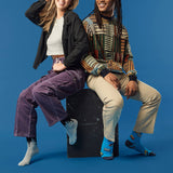 Two people sitting on a black box against a blue background wearing paint drip socks and cassette tape socks.