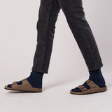 Person wearing brown sandals with blue socks and dark jeans on a light gray background thumbnail