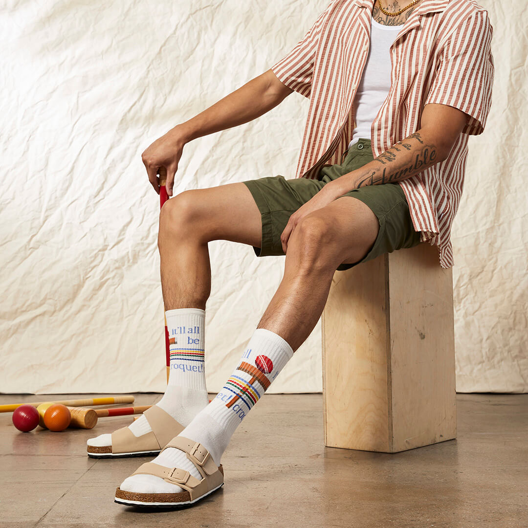 Person sitting on a wooden block wearing striped socks with colorful patterns, surrounded by croquet equipment.