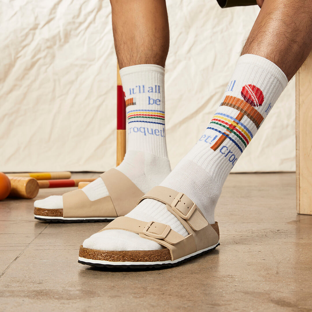 Person wearing white socks with colorful text and designs, paired with beige sandals on a wooden floor.