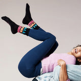 Person wearing colorful socks with text, blue leggings, and a pink sweater against a plain background
