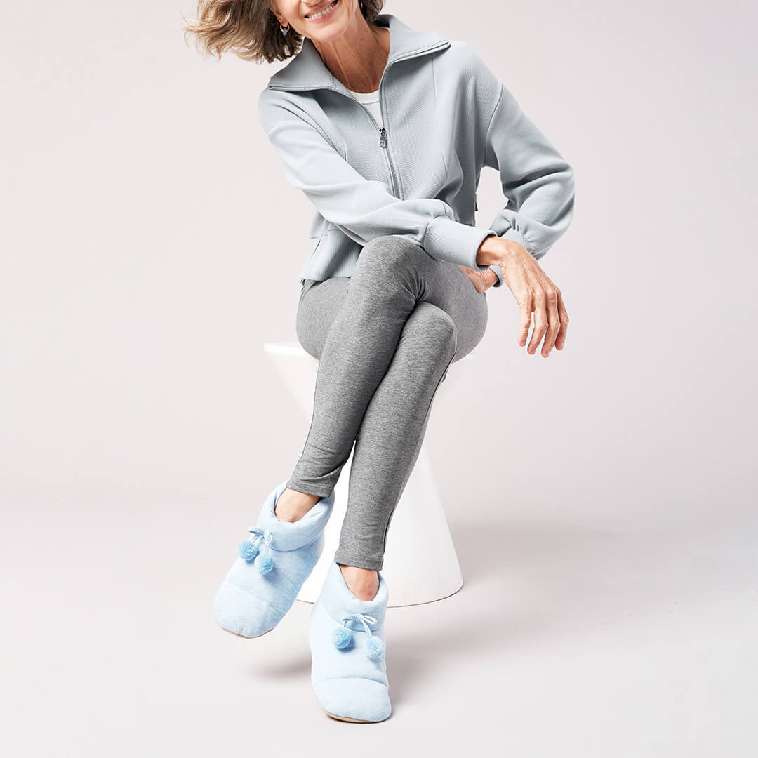 Person wearing a light gray tracksuit with blue jersey slippers on a white background