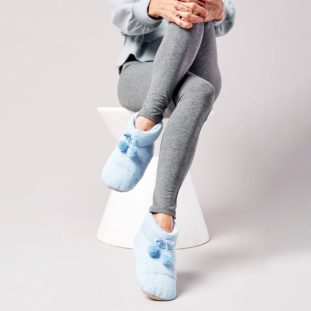 Person wearing light blue slippers with pom-poms sitting on a white stool against a plain background