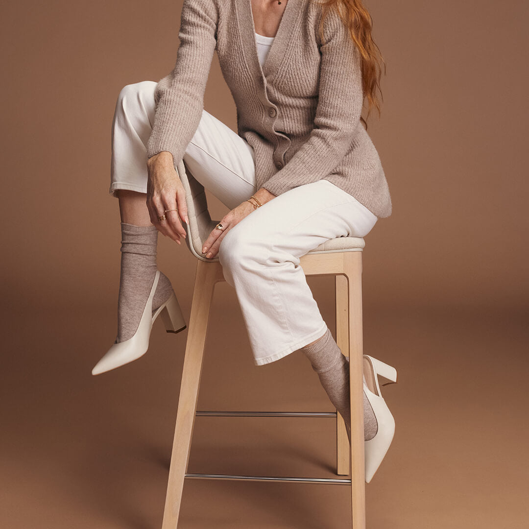 Woman sitting on a stool wearing a beige cardigan and socks, white pants, and high-heeled shoes against a brown background