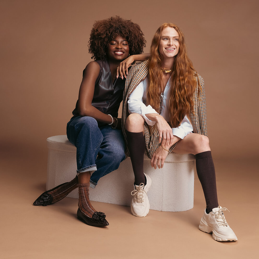 Two women sitting on a white stool against a brown background