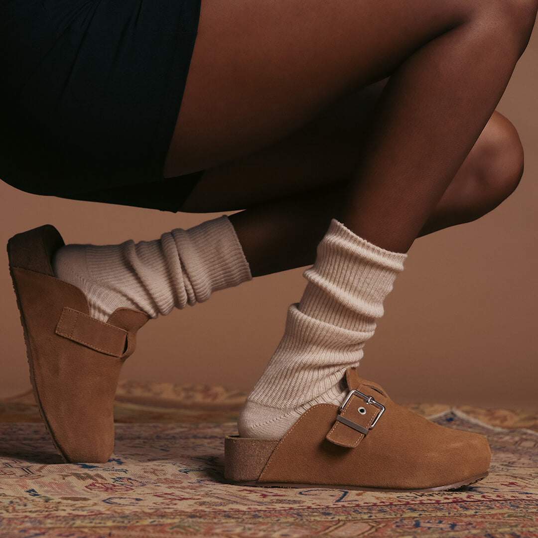 Brown suede clogs with buckles worn with beige slouch socks on a patterned rug.