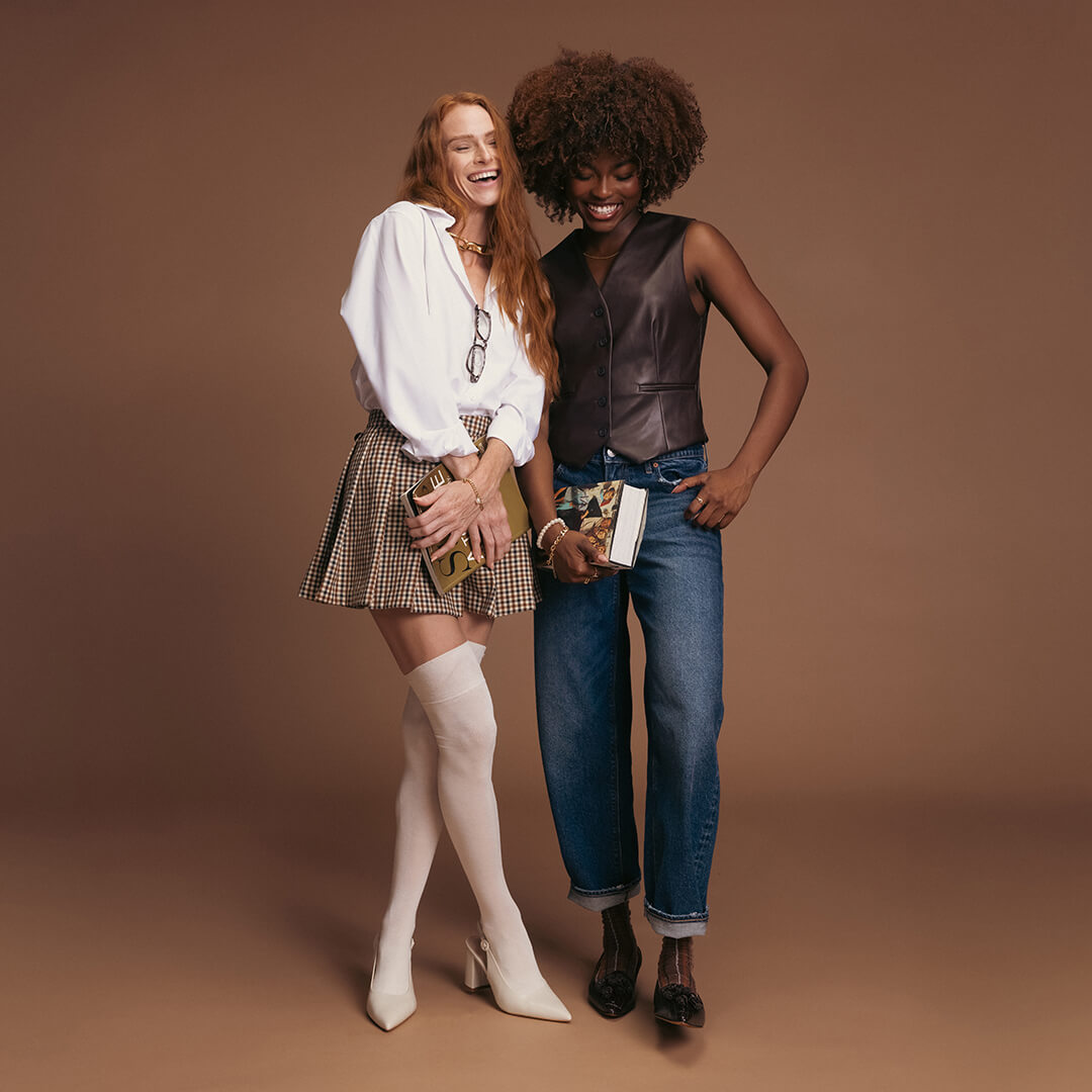 Two women standing together on a brown background, one wearing a white blouse and plaid skirt with white knee-high socks and high-heeled shoes, the other in a leather vest, jeans, and brown plaid-patterned socks with black pointed-toe shoes.